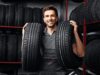 In a car repair shop, a man presents two tires that he is holding in his hands.