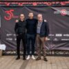 The Managing Director Mr. Strelen and Mr. Härle stand in front of a photo wall with Per Mertesacker. The photo wall is black.